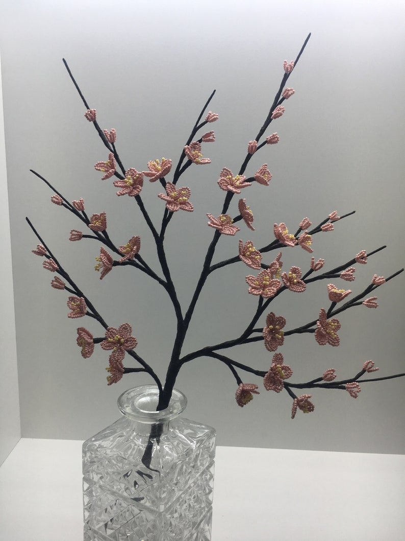 May include: A delicate pink beaded flower arrangement in a clear glass vase. The flowers are arranged in a branch-like shape, creating a realistic and elegant display.