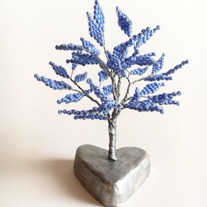 May include: A decorative tree sculpture featuring a silver wire trunk and branches adorned with small, light blue beads shaped like leaves. The tree is mounted on a solid, heart-shaped base with a metallic silver finish, creating a balanced structure.