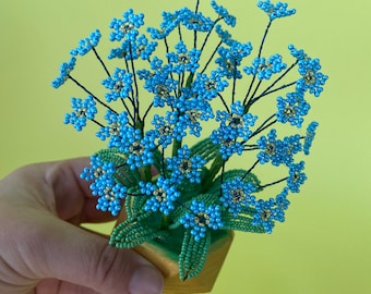 May include: A small potted plant made of blue and green beads. The plant is in a small wooden pot and has many delicate stems with blue flowers.