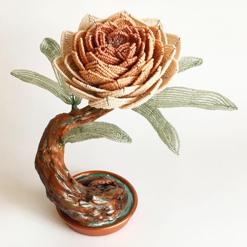 May include: A handcrafted beaded flower sculpture with a detailed, layered petal design in shades of beige and brown. The flower sits atop a curved, textured brown stem with green beaded leaves. The base is a copper-colored pot.
