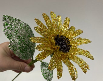 May include: A beaded sunflower with yellow petals and a brown center, with two green leaves. The sunflower is on a wire stem.
