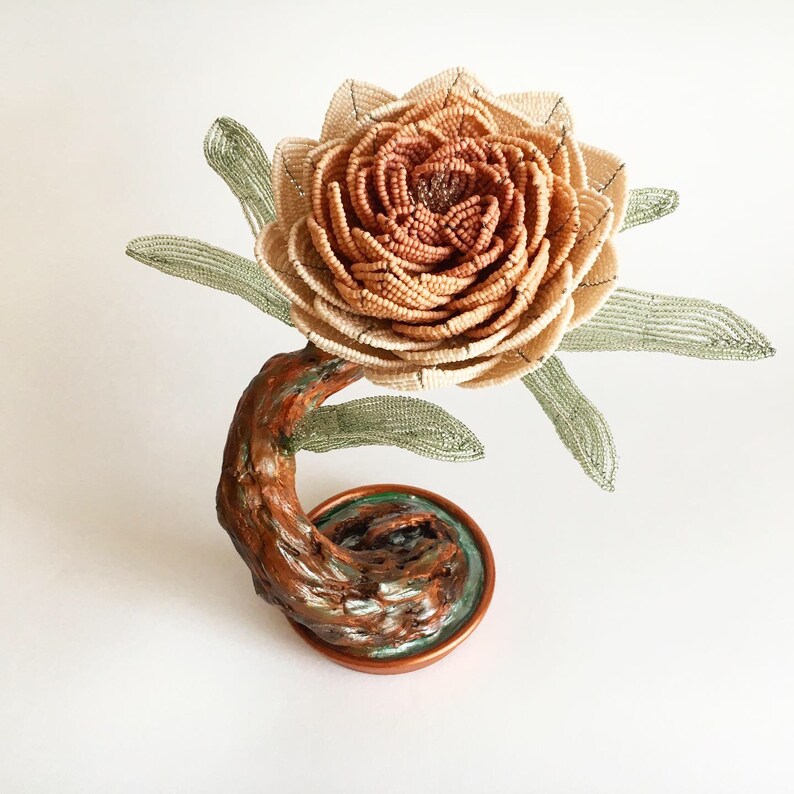 May include: A handcrafted, decorative flower sculpture. The flower features a layered, beige and brown beaded design, with green beaded leaves. The sculpture is supported by a brown, curved stem and a copper-colored base.