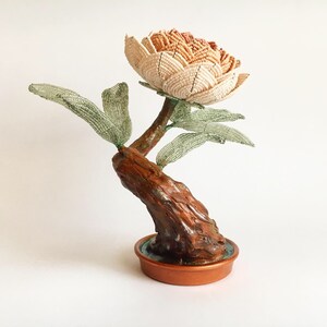 May include: A handcrafted beaded flower sculpture in a copper-colored pot. The flower has a beige and brown color scheme, with green beaded leaves and a brown, textured stem. The sculpture is a unique decorative item.