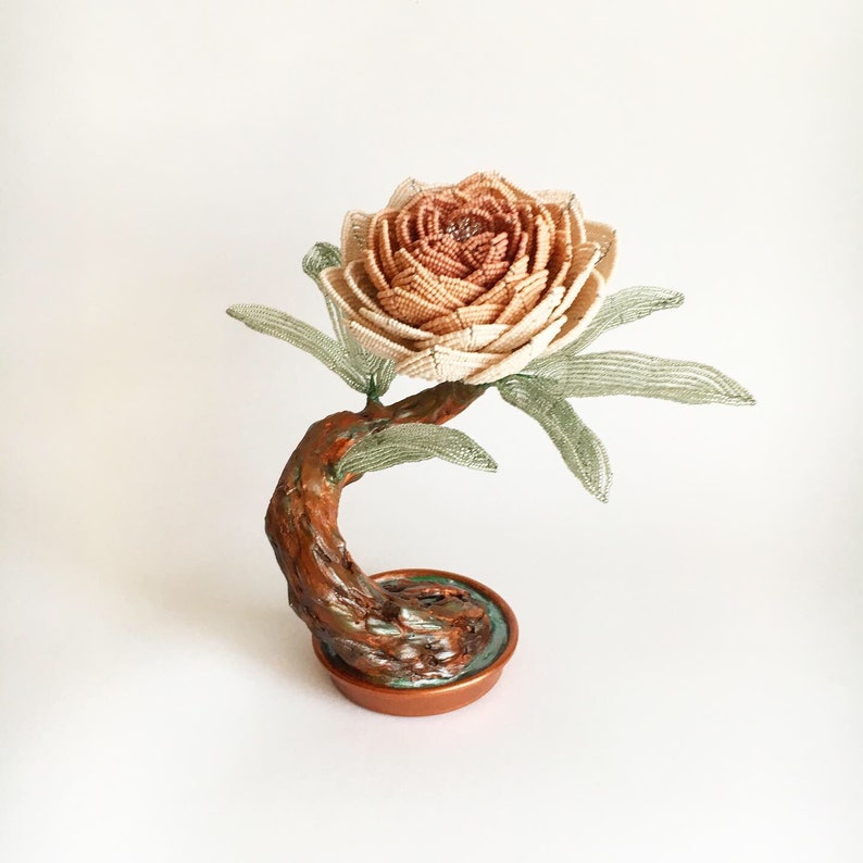 May include: A handcrafted beaded flower sculpture resembling a rose, with a gradient of beige to brown petals. The flower sits atop a brown, curved stem with green beaded leaves. The base is a copper-colored pot.