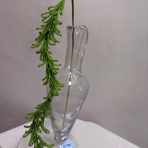 Könnte beinhalten: Ein dekorativer Perlenzweig aus kleinen, leuchtend grünen Glasperlen, die in einer langen, herabhängenden, kiefernartigen Form angeordnet sind, neben einer klaren, schlanken Glasvase mit Henkel und einem dezenten floralen Ätzmuster am Körper.