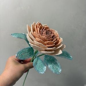 May include: A handcrafted flower sculpture made from small beads. The petals transition from a deep brownish-orange at the center to a pale cream at the outer edges. The flower features several bright teal green leaves attached to a thin stem.