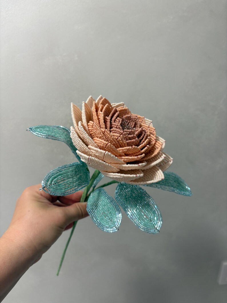 May include: A handcrafted flower sculpture made from small beads. The petals transition from a deep brownish-orange at the center to a pale cream at the outer edges. The flower features several bright teal green leaves attached to a thin stem.