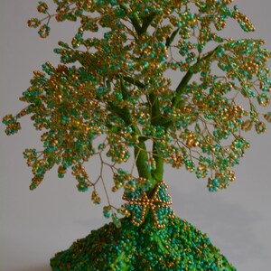 K&ouml;nnte beinhalten: Eine Perlenbaumskulptur mit einem gr&uuml;n-goldenen Farbschema. Der Baum besteht aus Draht und Perlen und hat einen kleinen Schmetterling an der Basis. Der Baum steht auf einem gr&uuml;nen Sockel.