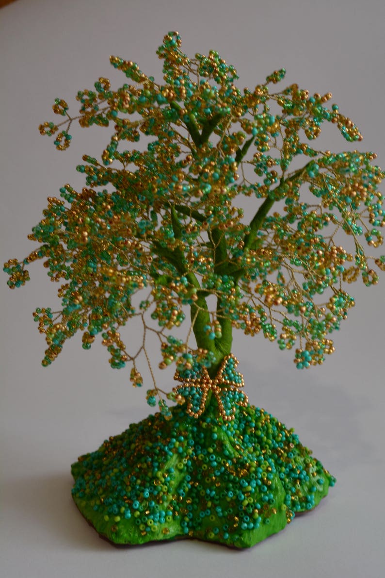 K&ouml;nnte beinhalten: Eine Perlenbaumskulptur mit einem gr&uuml;n-goldenen Farbschema. Der Baum besteht aus Draht und Perlen und hat einen kleinen Schmetterling an der Basis. Der Baum steht auf einem gr&uuml;nen Sockel.