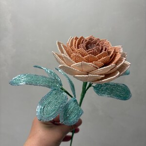 May include: A handcrafted beaded flower featuring a multi-layered bloom with petals transitioning from a deep peach center to a pale cream outer edge. The stem includes several light blue, translucent beaded leaves attached to a thin green wire.
