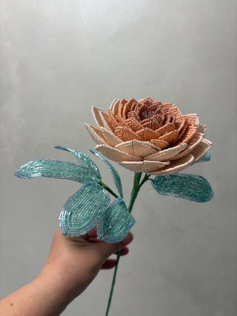 May include: A handcrafted beaded flower featuring a multi-layered bloom with petals transitioning from a deep peach center to a pale cream outer edge. The stem includes several light blue, translucent beaded leaves attached to a thin green wire.