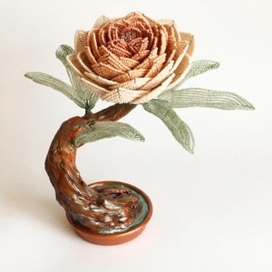 May include: A handcrafted beaded flower sculpture. The flower is a mix of beige and peach beads, with green beaded leaves. The stem is brown and the base is a copper-colored pot. A unique decorative piece.