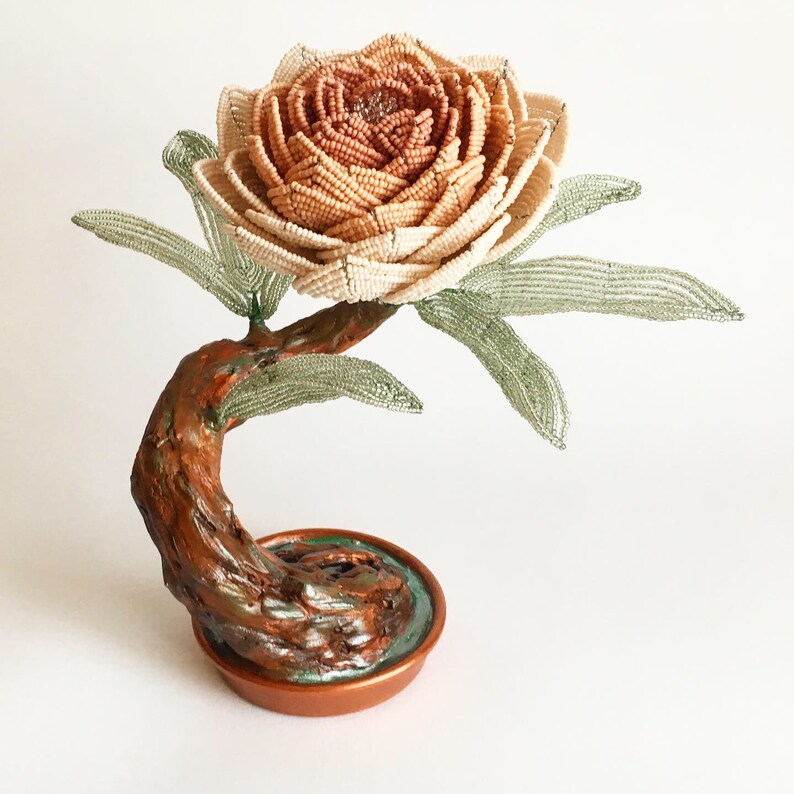 May include: A handcrafted beaded flower sculpture. The flower is a mix of beige and peach beads, with green beaded leaves. The stem is brown and the base is a copper-colored pot. A unique decorative piece.