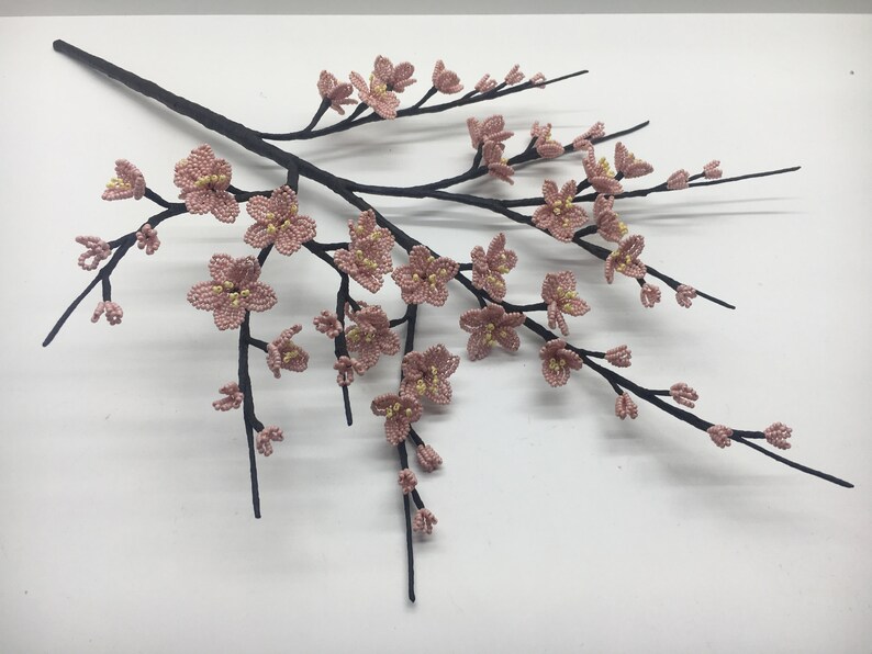 May include: A delicate branch with pink beaded blossoms, resembling a cherry blossom tree. The branch is made of black wire and the flowers are crafted with small pink and yellow beads.
