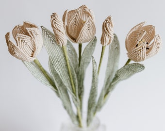May include: A bouquet of five cream-colored beaded tulips in a clear glass vase. The tulips are made of small white beads and have a delicate, feminine look.