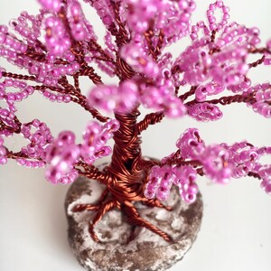 May include: A pink beaded wire tree with a brown wire base and a white base. The tree is made of small pink beads strung on wire and is shaped like a tree with branches.