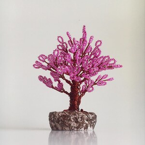 May include: A pink beaded wire bonsai tree with a brown wire trunk and a gray stone base.
