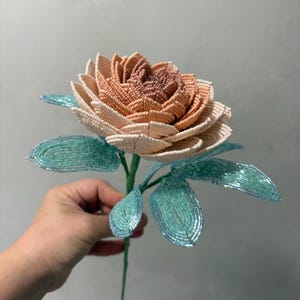 May include: A handcrafted beaded flower featuring a multi-layered bloom with petals transitioning from a deep dusty rose center to lighter cream-colored outer edges. The stem and pointed leaves are constructed from shimmering light turquoise beads.