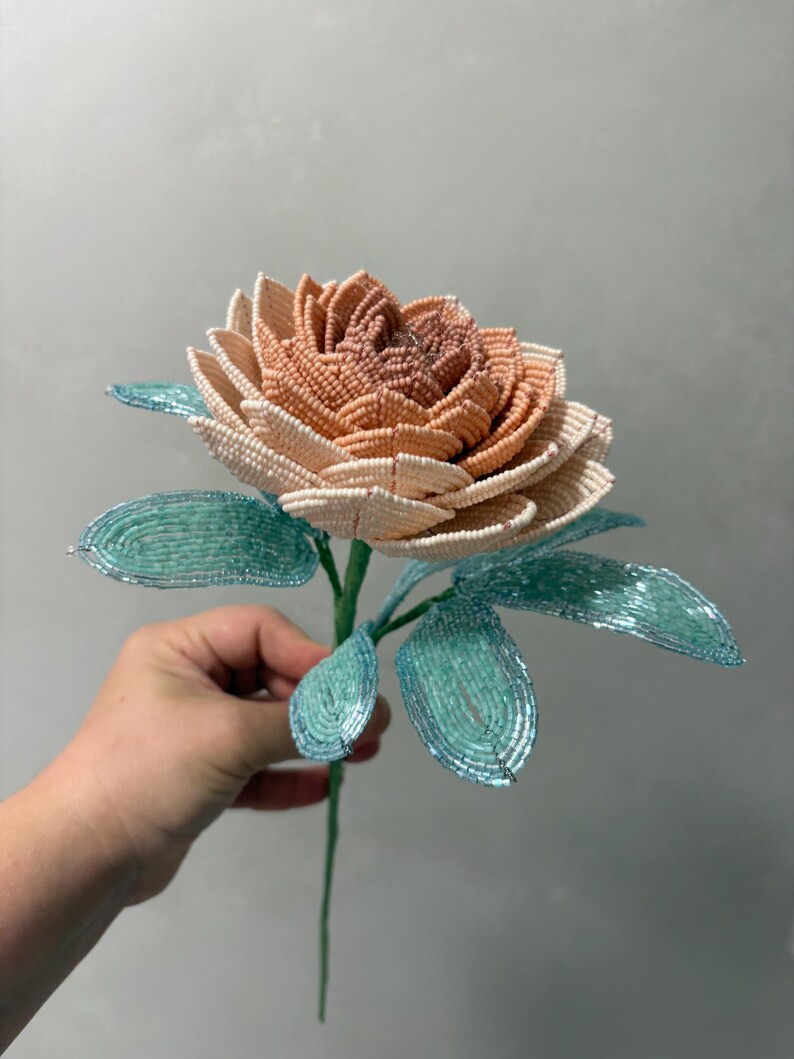 May include: A handcrafted beaded flower featuring a multi-layered bloom with petals transitioning from a deep dusty rose center to lighter cream-colored outer edges. The stem and pointed leaves are constructed from shimmering light turquoise beads.
