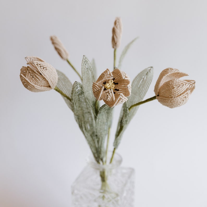 May include: A bouquet of beige beaded tulips with green beaded leaves in a clear glass vase.