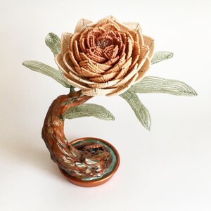 May include: A handcrafted beaded flower sculpture with a detailed, layered beige and brown flower head. The flower sits atop a curved, textured brown stem with green leaves. The base is a copper-colored dish.