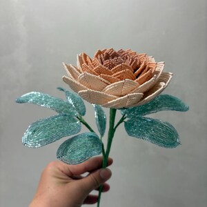 May include: A handcrafted beaded flower featuring a multi-layered bloom with petals transitioning from a deep peach center to light cream outer edges. The stem includes several textured, light blue-green leaves, all constructed from small glass beads.