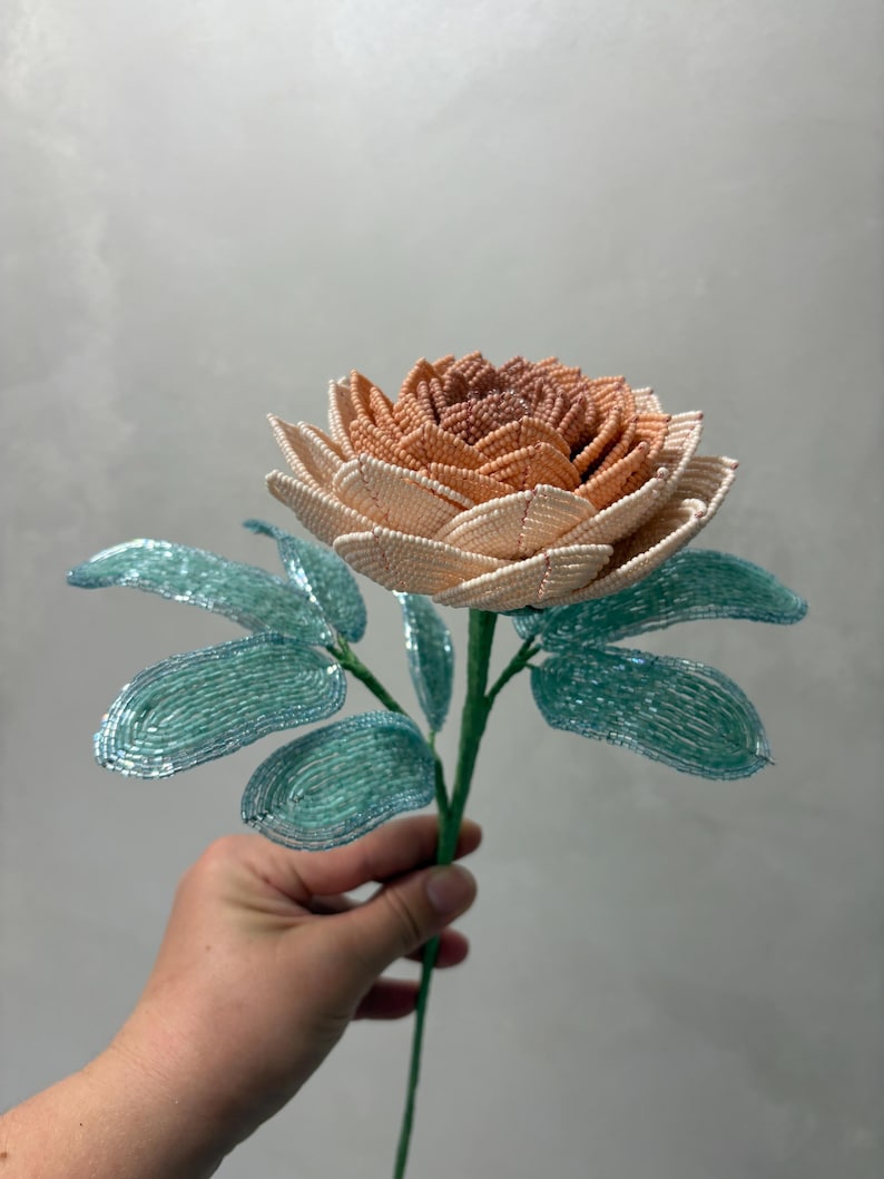 May include: A handcrafted beaded flower featuring a multi-layered bloom with petals transitioning from a deep peach center to light cream outer edges. The stem includes several textured, light blue-green leaves, all constructed from small glass beads.
