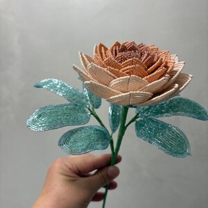May include: A handcrafted beaded flower featuring a multi-layered bloom with petals transitioning from a deep peach center to lighter cream edges. The stem includes several oval-shaped leaves made from shimmering light blue glass beads.