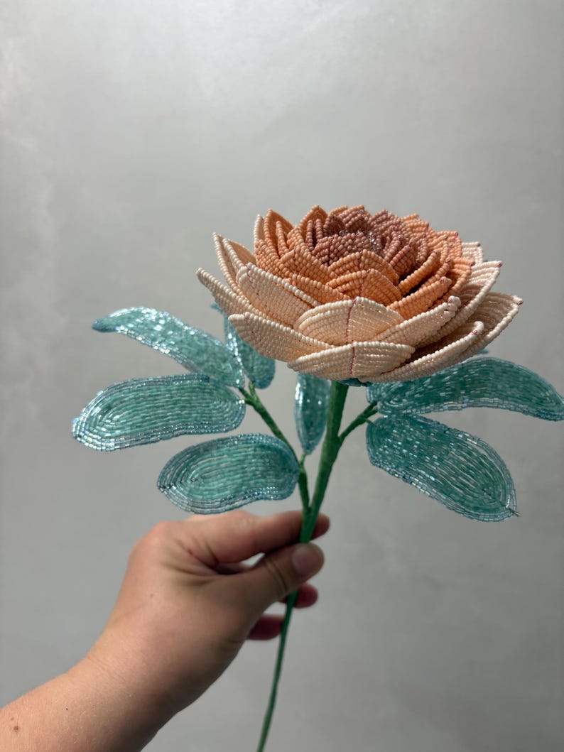 May include: A handcrafted beaded flower featuring a multi-layered bloom with petals transitioning from a deep peach center to lighter cream edges. The stem includes several oval-shaped leaves made from shimmering light blue glass beads.