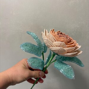 May include: A handcrafted flower sculpture made from small glass beads. The flower head features a gradient of cream, peach, and soft orange petals. It is attached to a green stem with several light blue, translucent beaded leaves.