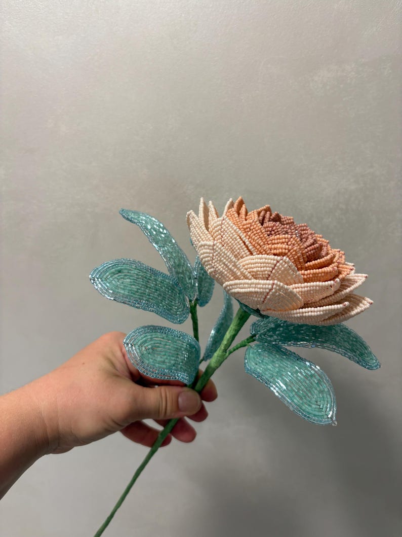 May include: A handcrafted flower sculpture made from small glass beads. The flower head features a gradient of cream, peach, and soft orange petals. It is attached to a green stem with several light blue, translucent beaded leaves.