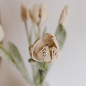 May include: A close-up of a beaded flower bouquet with a single white tulip in focus. The tulip is made of white beads and has a delicate, intricate design. The bouquet is a beautiful and unique piece of art.