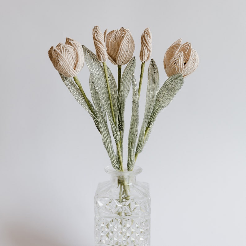 May include: A bouquet of five cream-colored beaded tulips with green beaded stems in a clear glass vase with a diamond pattern.