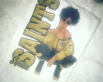 May include: A white t-shirt with a gold graphic of a woman wearing a yellow shirt and black pants. The graphic also includes the text "Saints Squad" and "Who Dat" with a New Orleans Saints logo.