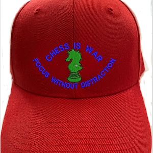 May include: A red baseball cap featuring an embroidered green chess knight piece in the center. The text CHESS IS WAR is arched above the knight, and the text FOCUS WITHOUT DISTRACTION is arched below, all stitched in a bright blue thread.