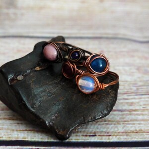 Op de afbeelding: Vier draadgewikkelde ringen met verschillende gekleurde edelstenen. De ringen zijn gestapeld op een donkergrijze steen. De edelstenen zijn roze, blauw, paars en wit.