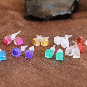 Könnte beinhalten: Eine Sammlung von quadratischen Ohrsteckern in verschiedenen Farben. Die Ohrringe sind in Blau, Lila, Grün, Klar, Rot und Gold erhältlich. Jeder Ohrring hat einen Stecker für durchbohrte Ohren. Die Ohrringe liegen auf einer braunen Oberfläche.