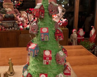 Könnte beinhalten: Ein grüner Papiermaché-Weihnachtsbaum mit einem Stern oben. Der Baum ist mit kleinen Häusern geschmückt, die Nummern tragen. Die Häuser sind rot, blau und grün. Der Baum steht auf einem Holztisch.