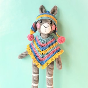 Op de afbeelding: Een grijze gehaakte bunny die een kleurrijke gestreepte poncho draagt en een gele en blauwe gehaakte muts met pompoenen. De bunny heeft een wit gezicht met zwarte ogen en een roze neus. De bunny staat op een lichtblauwe achtergrond. "Mrs. Milly" staat in roze op een witte wolk.