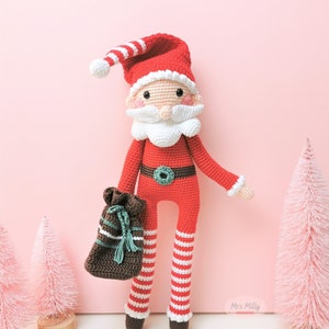 Op de afbeelding: Een gehaakte kerstmanpop met een witte baard, rood-wit gestreepte benen en een bruine zak in zijn hand. De pop staat op een roze achtergrond met twee kleine roze bomen.