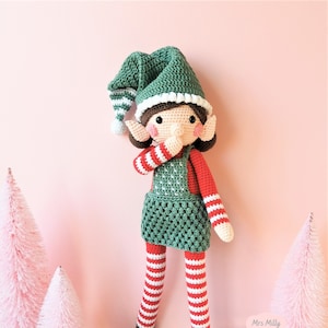 Puede incluir: Una muñeca de elfo de crochet con un gorro verde, una camisa y leggings a rayas rojas y blancas y una falda verde. La muñeca está de pie sobre un fondo rosa con dos pequeños árboles rosas.