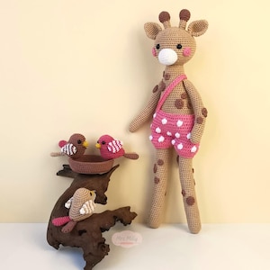 Op de afbeelding: Een gehaakt giraffen speelgoed dat een roze broek met witte stippen draagt, staat naast een houten tak met drie gehaakte vogelspeelgoed. De vogels zijn bruin, wit en roze.