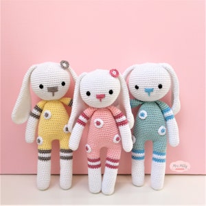 Op de afbeelding: Drie gehaakte konijnen speelgoed met verschillende gekleurde outfits. Het konijn links draagt een gele outfit met bruine strepen, het konijn in het midden draagt een roze outfit met rode strepen en het konijn rechts draagt een blauwe outfit met blauwe strepen.