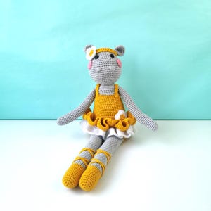 May include: A handmade gray crochet hippo doll wearing a yellow dress with a white ruffled skirt and matching yellow ballet slippers. The doll has a flower headband and pink cheeks, perfect for a child's toy or nursery decor.