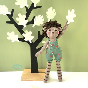 Peut inclure: Un hérisson en crochet marron et beige portant une salopette verte avec des accents de champignons rouges. Le hérisson est debout sur une base en bois avec un arbre noir derrière lui.