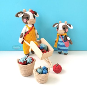 Op de afbeelding: Twee gehaakte koeienpoppen met bruine en witte vlekken, roze wangen en hoorns. De een draagt een gele tuinbroek, de ander een blauw gestreepte tuinbroek met een gele sjaal. Een houten kruiwagen bevat kleine gehaakte vruchten bij twee mandjes.