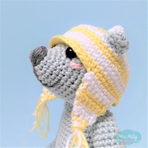 Op de afbeelding: Een grijs gehaakt knuffeldier met een gele en witte gestreepte muts met een bijpassende sjaal. Het dier heeft een kleine, grijze neus en zwarte ogen.