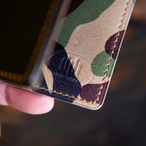 Op de afbeelding: Close-up van een leren portemonnee met camouflagepatroon. De portemonnee heeft een mix van groene, bruine en paarse tinten. De randen zijn gestikt met gouden draad. Het reliëflogo luidt "HIGH GRAIN".