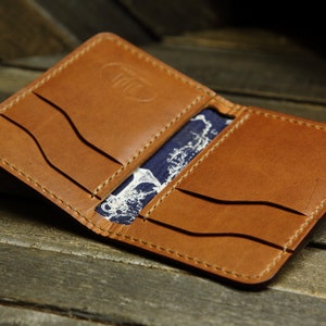 Op de afbeelding: Open, bruinleren portemonnee met zichtbare stiksels en meerdere kaartsleuven. De binnenkant heeft een blauw-witte stof met patroon. De portemonnee wordt open getoond, waarbij het ontwerp en de afwerking worden getoond.