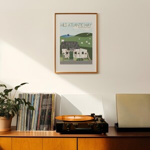 May include: A framed print of a cottage with a green door and a green roof. The print is titled "Wild Atlantic Way, Ireland's West Coast". The print is hanging on a white wall above a record player and a stack of vinyl records.